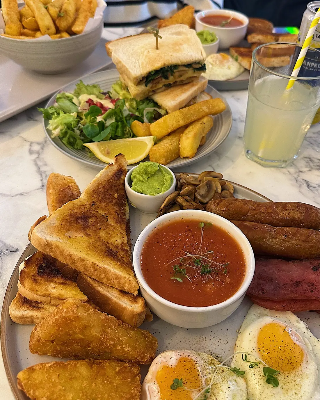 A full table at TenTen from above — a full English with toast and hash browns, a stacked club sandwich with halloumi fries, a bowl of chips and a fresh lemonade on marble.